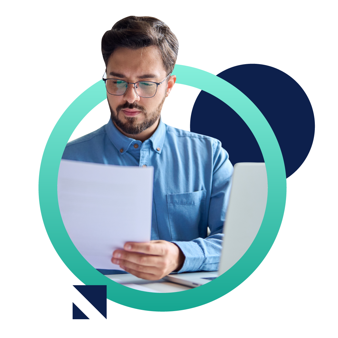 A business man in a blue buttoned shirt looks at a document while sitting at his desk in front of a computer, all placed inside of a designed green circle.