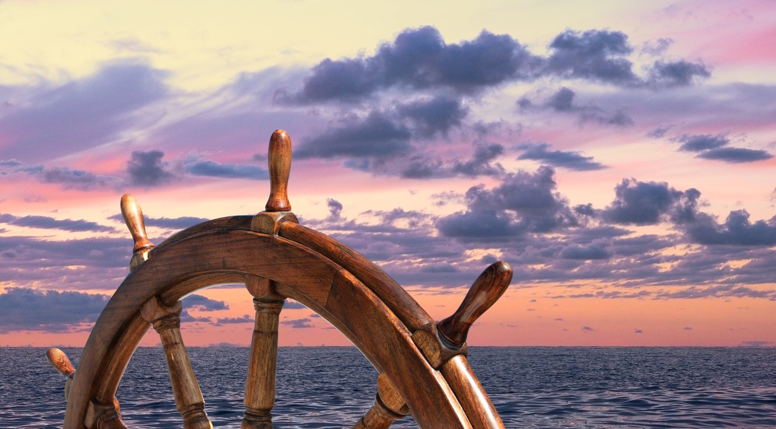 A ships steering wheel in the foreground overlooking calm seas at a sunset.