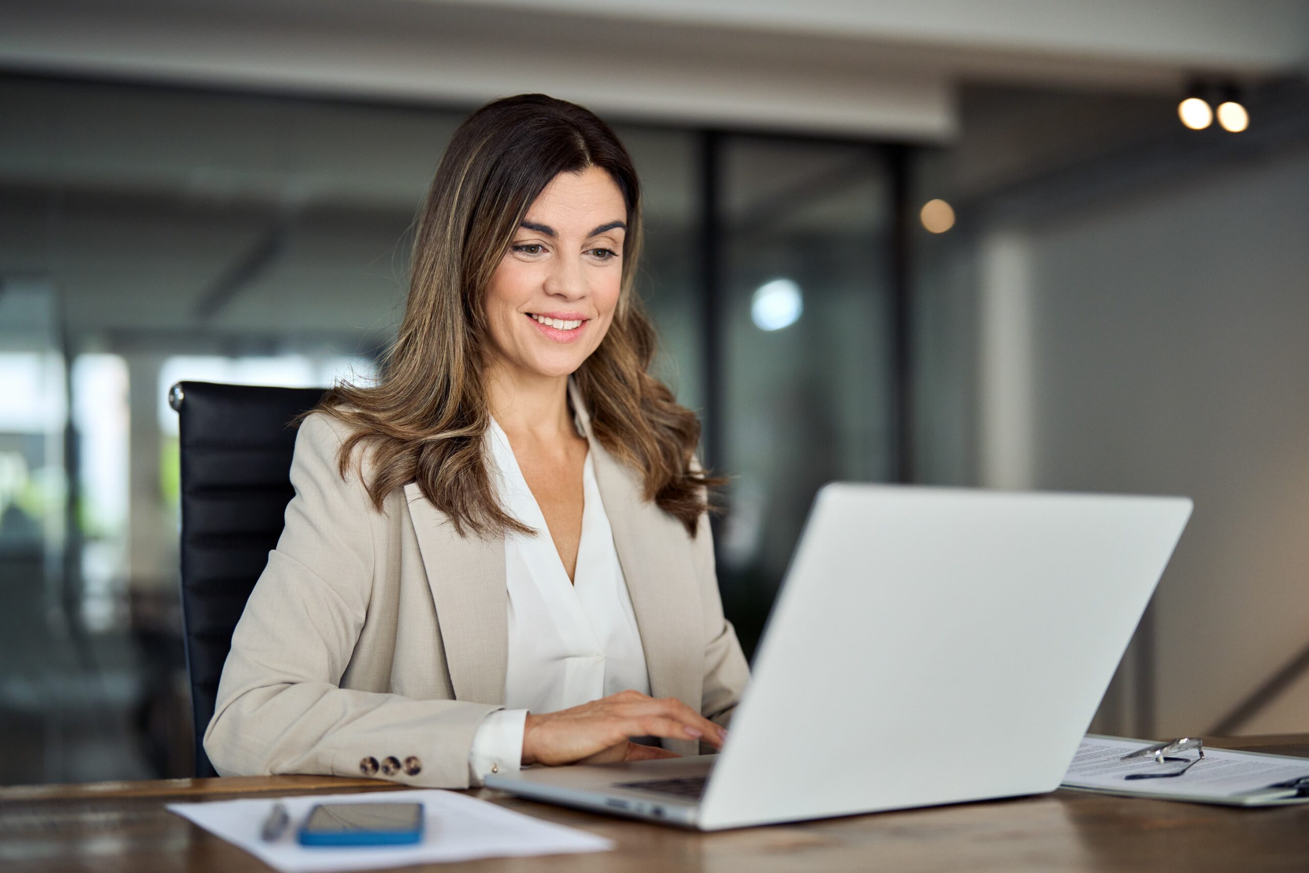 shutterstock_2353012815-min-scaled A professional business woman sitting at a table and typing on her laptop.