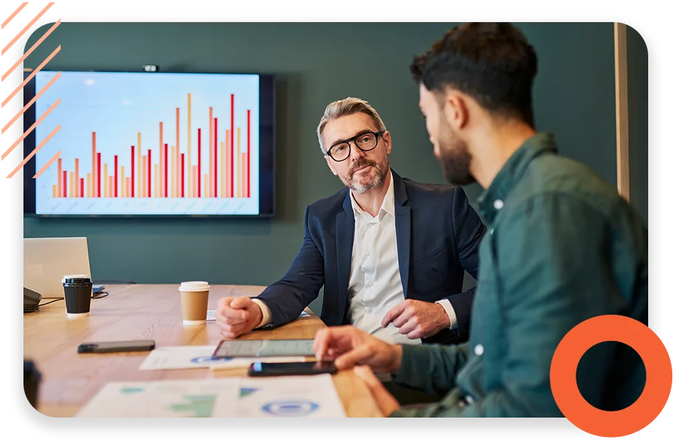Two business men in a conference room working and talking with an upward chart in the background.