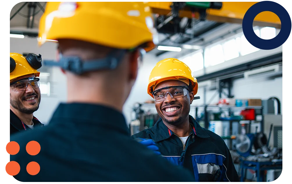 Three manufacturing colleagues smiling in a factory while wearing hard hats.